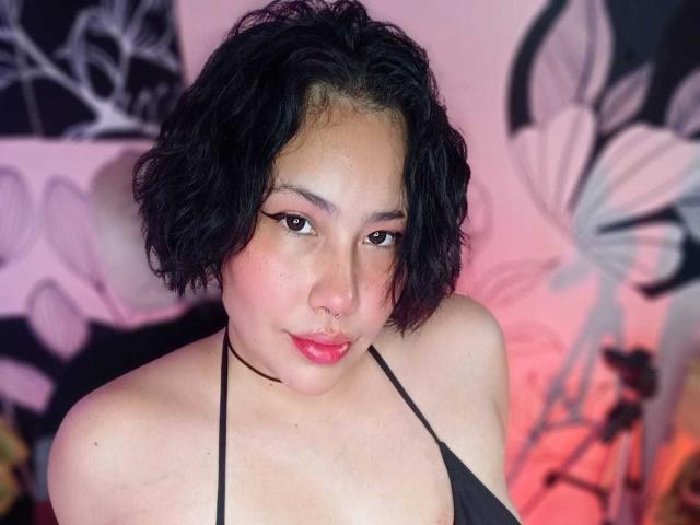 RebbekaBhons, con cabello negro rizado corto, un aro en la nariz, sombra de ojos rosa, un top halter negro y lápiz labial rosa, mostrando sus senos y un rostro adorable, sobre un fondo iluminado en rosa..