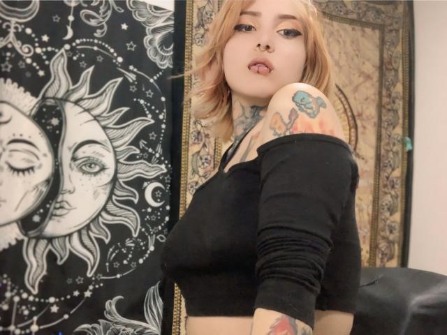 AliceCalloway with short blonde hair, black off-shoulder top, tattoos on her right arm and neck, nose ring, and tongue piercing, showing her tits and looking at the camera.