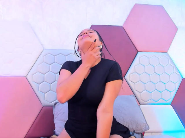Arielle Wilde, mit der Hand am Hals, die Augen geschlossen, ein schwarzes Hemd tragend, sitzt an einer Wand mit sechseckigem Muster und wirkt gestresst oder hat Schmerzen. Ihr Hintern ist sichtbar..
