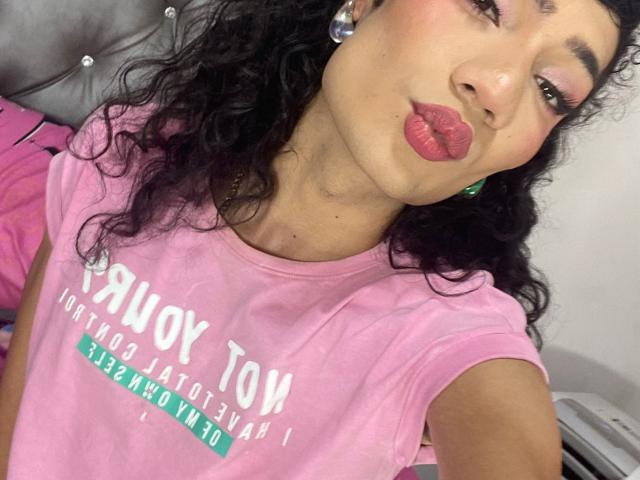 SweetSamyPrincipal has curly black hair, pink lipstick, and is pouting her full, round ass-lookin' lips. She's wearing a pink shirt and green earrings. Her skin is tan.