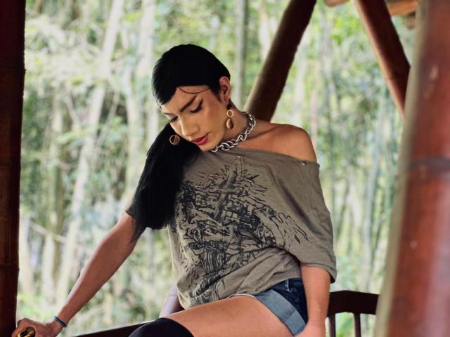 SweetSamyPrincipal with black hair, off-shoulder grey tee, chain necklace, denim shorts, sitting on wooden chair, looking down, wearing large gold earrings, forest background.
