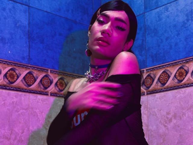 SweetSamyPrincipal with short black hair, wearing a black off-shoulder top, blue choker, and large earrings, covering their chest with a blushing, pink-lit, slightly blurred, sultry expression.