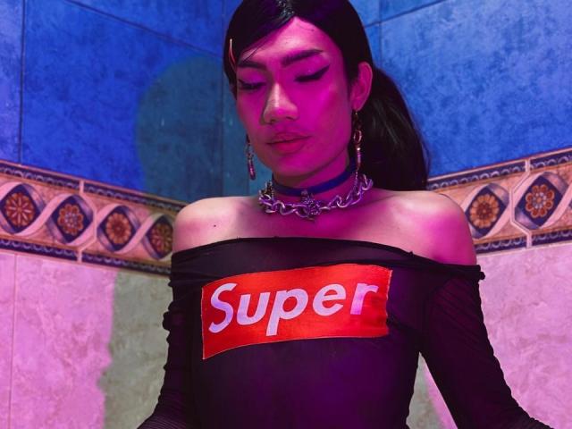 SweetSamyPrincipal with dark hair, wearing a black off-shoulder top with "Super" in red, chain choker, pink-blue lighting, and eyes closed, in a tiled bathroom.