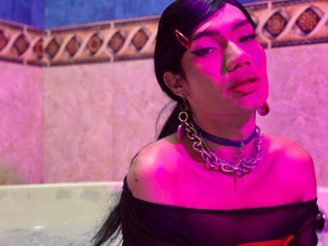 SweetSamyPrincipal with pink-tinted skin, black sheer top, multiple metal chains, blue choker, black hair in a ponytail, pink hairclip, and earrings, looking off to the side.