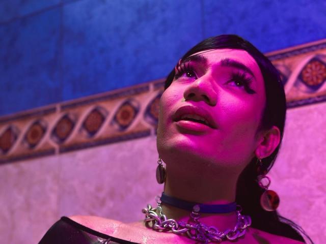 SweetSamyPrincipal has pink-tinted skin, dark hair, and thick eyelashes, wearing a chain choker and earrings, looking up with a slightly open mouth, bathed in purple and blue light.