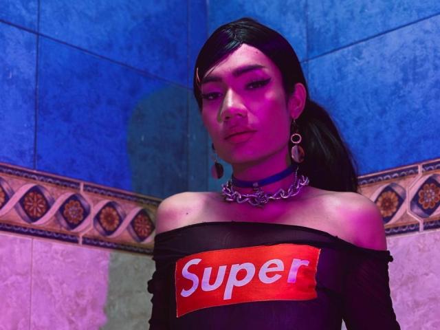 SweetSamyPrincipal with dark hair in a ponytail, wearing a black off-shoulder top with "Super" text, pink and blue makeup, chain necklace, and purple lighting, looking at the camera.
