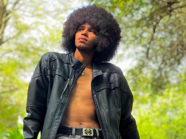 AntonyCarterr with a big afro, wearing a black leather jacket, shirt open to show a bare chest, standing outdoors, looking up with a serious expression.