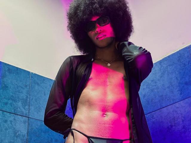 AntonyCarterr with afro, glasses, black open jacket, showing a toned, pink-lit chest and wearing black underwear, has a small necklace and a confident, slightly smug expression.