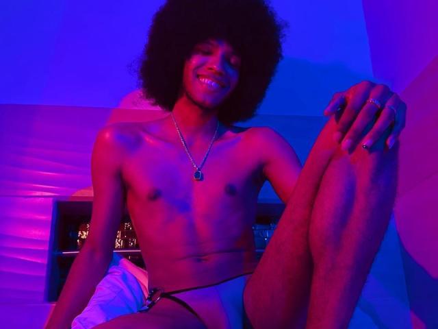 AntonyCarterr, topless, with an afro, wearing only black and white jockstrap, sitting with legs up, smiling, blue and pink lighting, wearing necklace with black pendant, hands on knees.