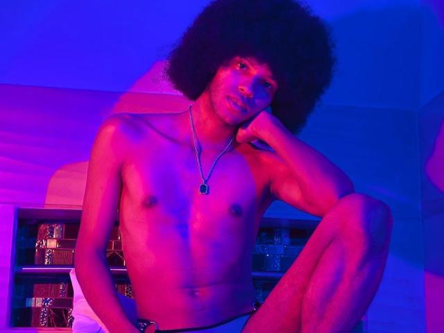 AntonyCarterr with a big afro, shirtless, wearing white underwear, blue and pink neon lights, sitting with one knee up, hand on chin, necklace, looking at camera.