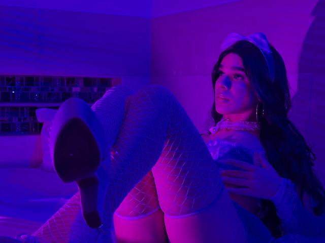 KataHot69 with blue-pink lit skin, wearing fishnet stockings, white high heels, and a cat ear headband, sitting with legs up, hand on her tits, looking seductively.