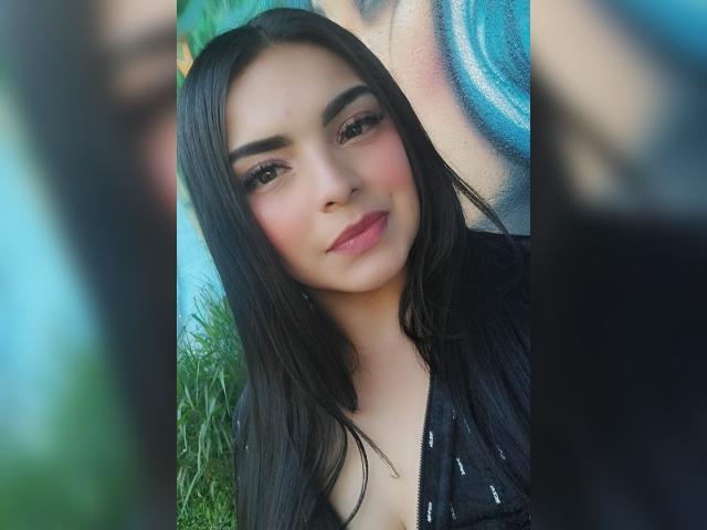 CarlaFiorell tiene el pelo largo y negro, ojos marrones, rubor rosado y pintalabios rosa. Lleva una blusa negra, brillante y con cremallera. Se puede ver la escote. Sonríe ligeramente y está al aire libre..