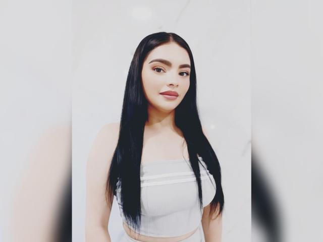CarlaFiorell with long black hair, wearing a white strapless top, showing her small tits, smiling at the camera, light skin, and subtle makeup, against a plain white background.