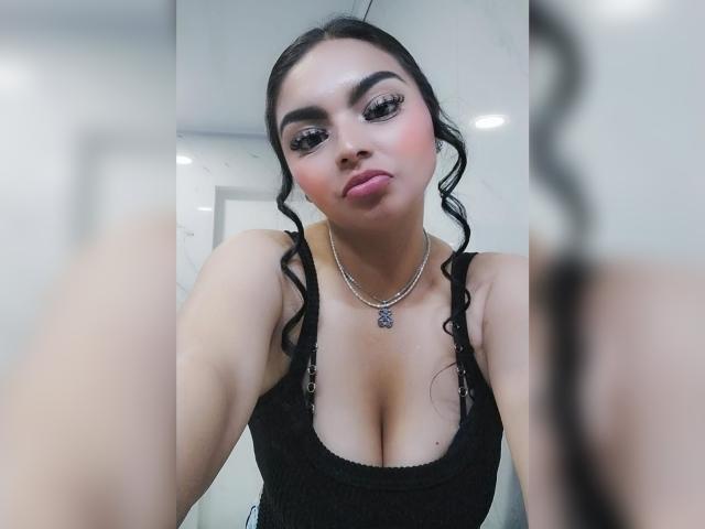 CarlaFiorell with wavy black hair, thick eyebrows, long eyelashes, pink lipstick, black tank top showing her big tits, silver necklace, and a pouty face in a selfie.