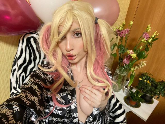 ElzaKomx with blonde pigtails, pink tips, zebra-pattern robe, black and white text robe, hand on chest, pouty lips, pink and white balloons, flowers, and potted plants in background.