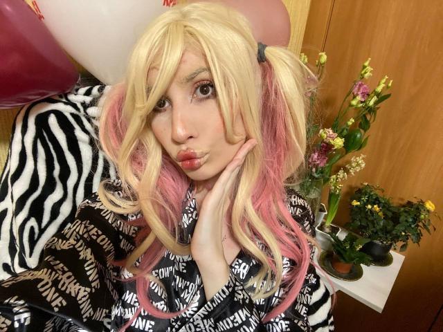 ElzaKomx with blonde and pink pigtails, zebra-patterned shirt, pouty lips, and hand on cheek, sitting near balloons and flowers, showing off her cute and sexy face.
