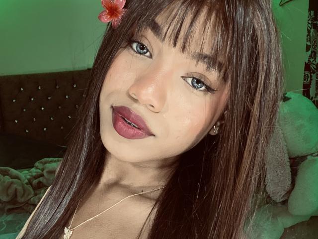 KathaThoms has straight brown hair with a red flower, dark eyeliner, pink lipstick, and a gold necklace. She's smiling with green lighting in the background.