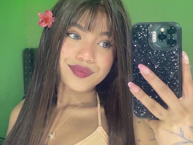 KathaThoms with long brown hair, pink flower, and pink lipstick, holding a black phone with a speckled case, smiling, showing her tits in a mirror selfie.