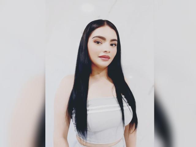 CarlaFiorell with long black hair, wearing a white strapless crop top, showing her tits, standing against a white background, smiling with red lipstick.