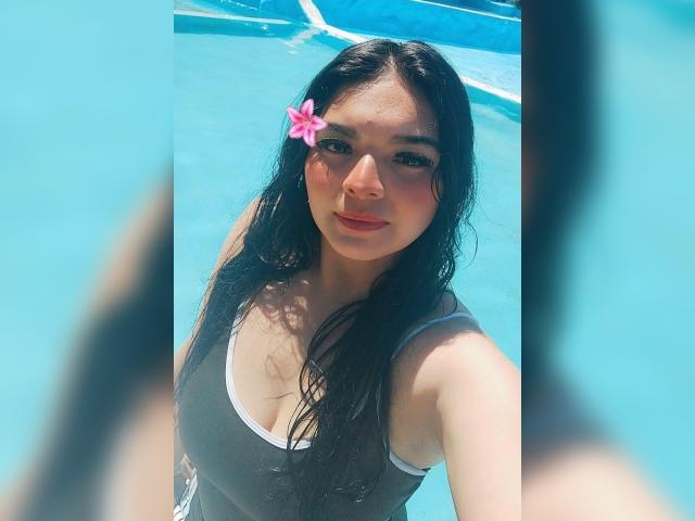 CarlaFiorell con i capelli neri bagnati, un fiore rosa tra i capelli, indossa un costume intero grigio, acqua blu, guarda verso l'alto, seni visibili, carina, leggermente arrossata, giornata di sole, selfie in piscina..
