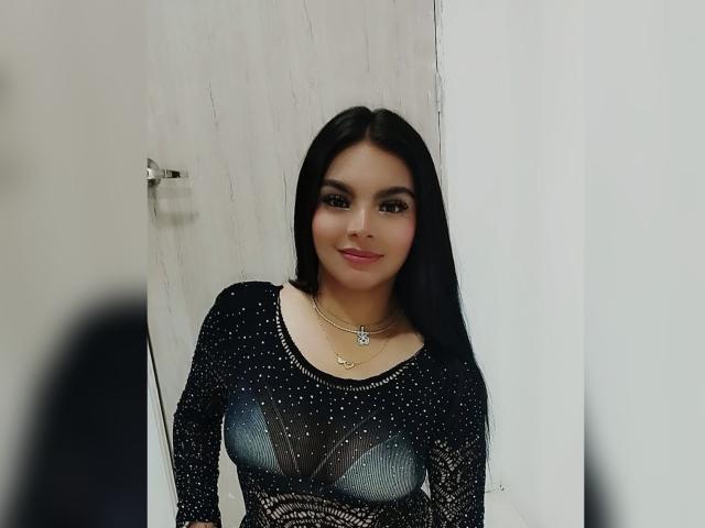 CarlaFiorell has long black hair, wearing a sparkly black top with glittery patterns, showing her boobs, and a necklace with a small pendant, smiling slightly.
