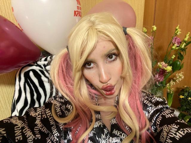 ElzaKomx with blonde and pink pigtails, pouty lips, wearing a zebra-patterned and black-and-white patterned top, with balloons and flowers in the background.