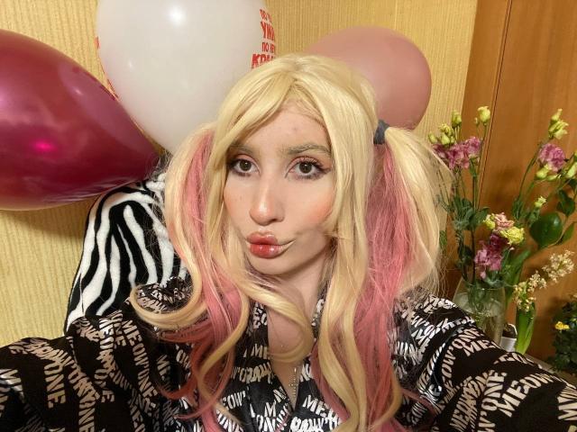 ElzaKomx with blonde and pink pigtails, pouty pink lips, wearing a zebra-patterned shirt, surrounded by balloons, making a kissing face. Her tits are not visible.
