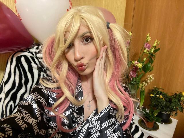ElzaKomx with blonde and pink pigtails, pouty lips, and large brown eyes, wearing a zebra-patterned shirt, touching her face. Balloons and flowers in the background.