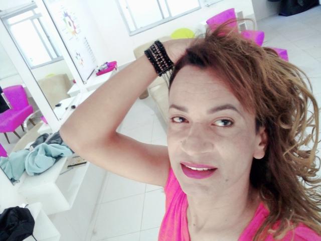 BrunelaSwan with wavy brown hair, pink shirt, black bracelets, smiling, hand in hair, pink lipstick, visible small mole on right cheek, white tiled room, pink chair in background.