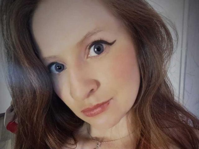 EnzumGinger has blue eyes with thick black eyeliner, light brown wavy hair, and pink lipstick. She's wearing a delicate necklace, smiling softly at the camera.