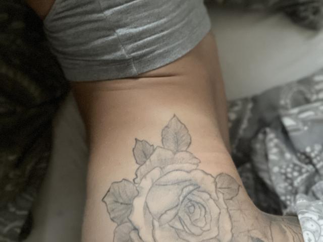 Le bras tatoué de DaneeSoHot, avec une rose détaillée en noir et gris sur l'avant-bras, portant une chemise grise, montrant un léger ventre au-dessus du tatouage..