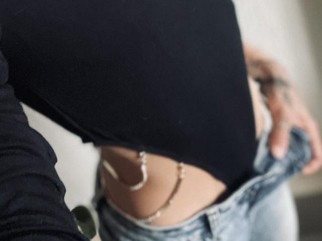 DaneeSoHots schwarzes Oberteil ist hochgezogen, wodurch eine Bauchnabelkette und ein Hauch ihres nackten, gebräunten Unterbauchs sichtbar werden. Sie öffnet die Knöpfe ihrer Jeans..