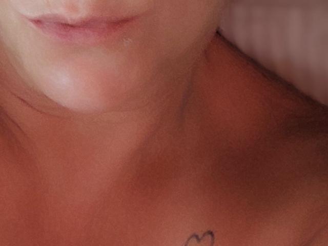 Line2323's upper chest and chin, with a small heart tattoo on her right shoulder, showing a slight, pinkish, relaxed smile and smooth, tan skin.