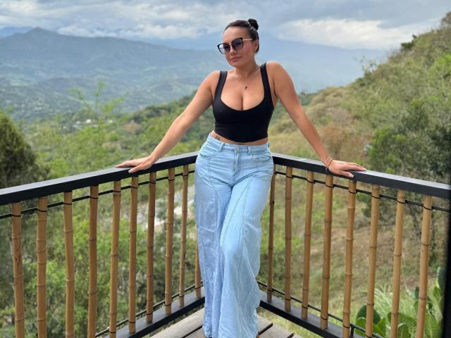 GalaMalkova with big tits, wearing a black tank top and light blue high-waisted pants, standing on a bamboo balcony, arms on rail, looking at camera, wearing sunglasses.