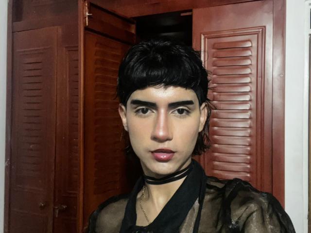 IvyMiller with dark, curly hair, brown eyes, and pink lips, wearing a sheer black top and black choker, standing in front of wooden doors.