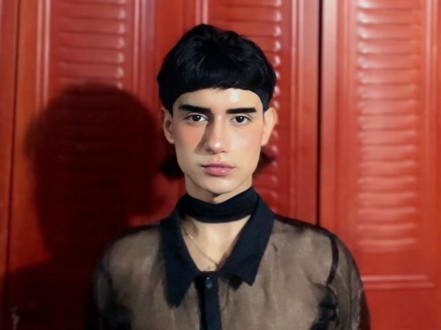IvyMiller with short black hair, wearing a sheer black top with a black choker, standing against red louvered doors, looking serious with a slightly pouty expression.