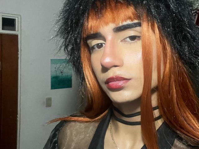 IvyMiller with orange and black hair, black furry hat, thick black eyebrows, red lipstick, black choker, and sheer black top, showing a serious face.