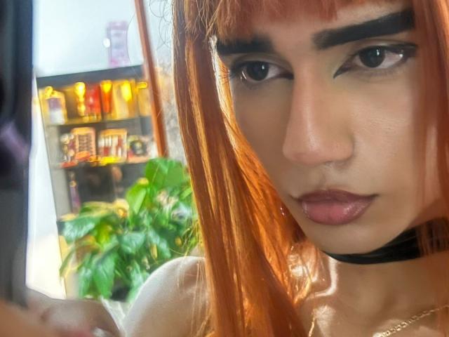 IvyMiller with red hair, thick eyebrows, dark eyeliner, and pink lipstick, wearing a black choker, holding a phone, looking serious, with a green plant in the background.