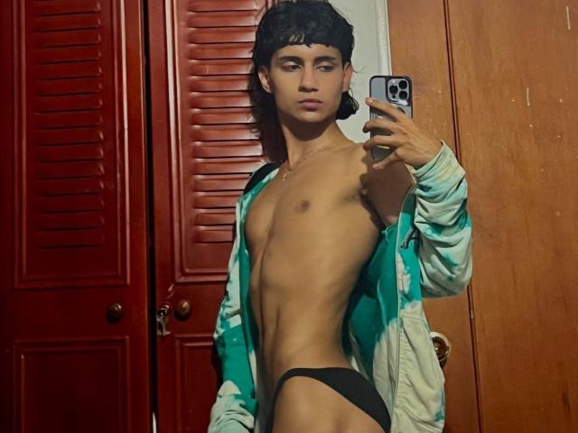 IvyMiller, with short black hair, wearing an open green jacket and black jockstrap, showing a toned, flat chest and visible penis, taking a mirror selfie with a smartphone.