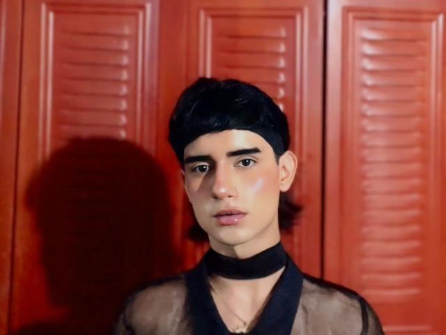 IvyMiller with black hair, black choker, and sheer black top, has a serious expression, standing in front of red louvered doors, with a shadow on the left.