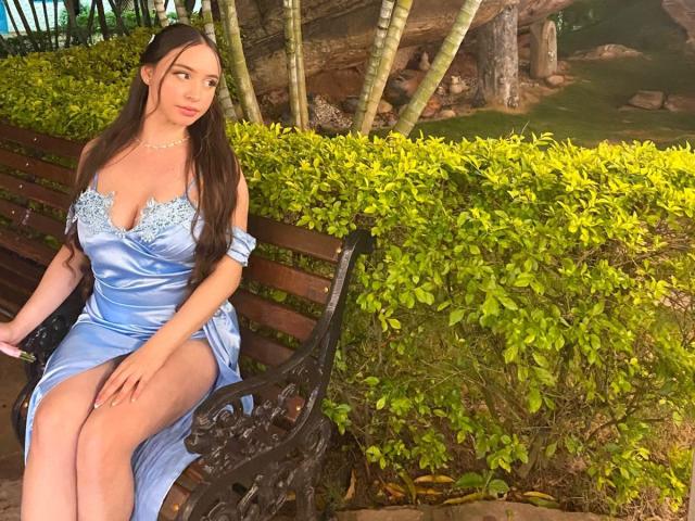 SophieHale with long brown hair, wearing a blue satin dress with lace trim, sitting on a black iron bench, showing her tits and a leg, in a sunny garden.