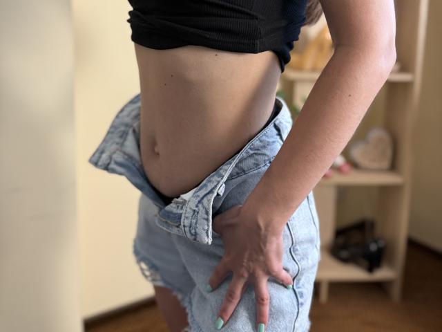 Stefanni's midriff with a black crop top, light blue denim shorts, and hand on her crotch. Her nails are green, and she's showing her flat, toned stomach.