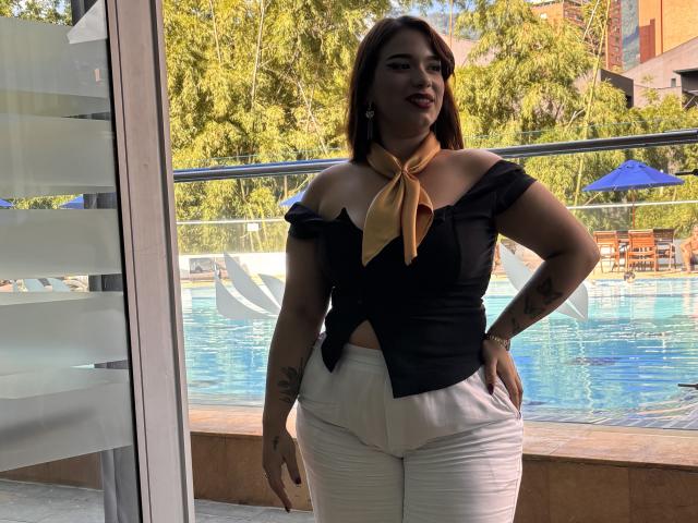 Milly Johnson, com cabelo castanho, blusa preta com um ombro à mostra, lenço bege, calças brancas, em pé perto de uma piscina, com a mão no quadril, tatuagens nos braços, olhando para a câmera, ao ar livre, ensolarado..