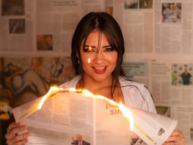 LizzeJoy, avec ses cheveux bruns foncés, tient un journal ouvert. Elle a des étincelles sur sa poitrine, elle sourit avec une lueur dans les yeux, elle porte une chemise blanche, sur une photo chaleureuse..