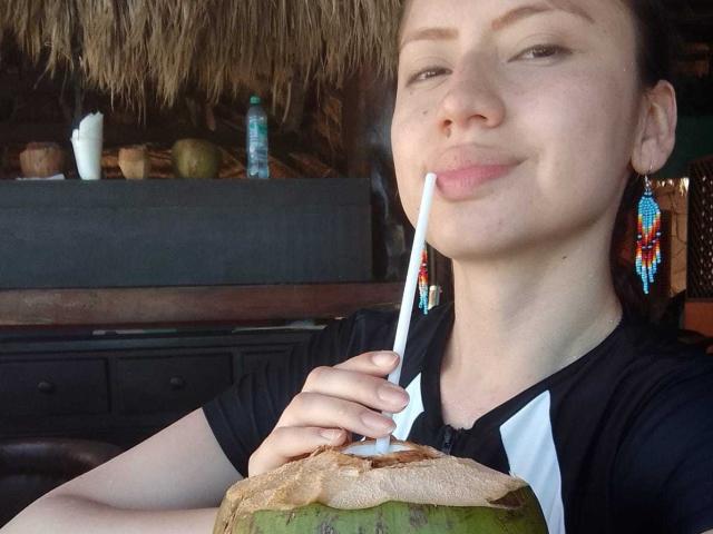 ElizaCanterville with a smirk, holding a green coconut with a straw in her mouth, wearing a black and white shirt, and colorful earrings.