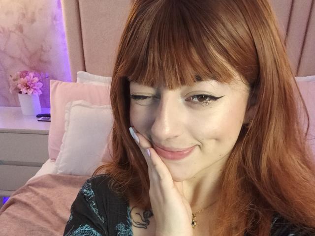 AnntonellaWatson with red hair, smiling, winking, hand on cheek, black eyeliner, pink lips, small tattoo on hand, gold necklace, wearing black top, on pink bed.