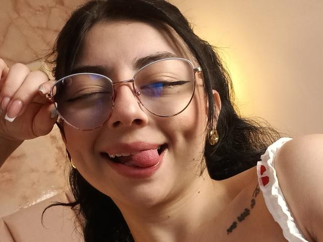 AnntonellaWatson with dark hair, glasses, tongue out, small gold earrings, white strap dress with red hearts, tattoo on right side of neck, holding glasses with right hand.