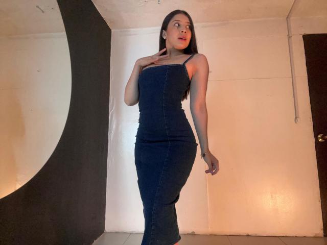 EmmaLombardi standing in a room, wearing a tight blue denim dress, her hand on her chest, showing her curvy ass and small tits, with long dark hair.