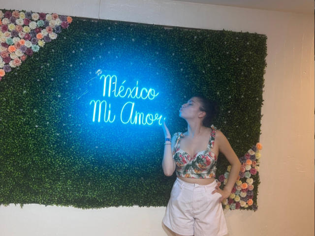 ElizaCanterville with floral crop top, showing tits, and white shorts, gazing at neon sign, hand on hip, green wall with flowers, blue neon light.