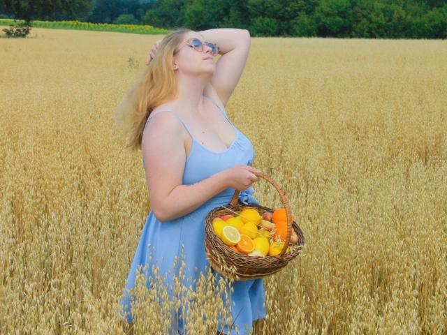 JessikaDaniels with blonde hair, blue dress, holding wicker basket of oranges, lemons, and carrots, standing in golden wheat field, arm raised, eyes closed, looking relaxed.